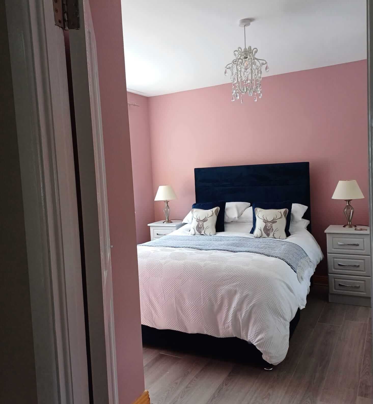 a bedroom with pink walls, a navy velvet bed and a glass style, small chandelier. Two white, wooden side tables with lampshades on each.